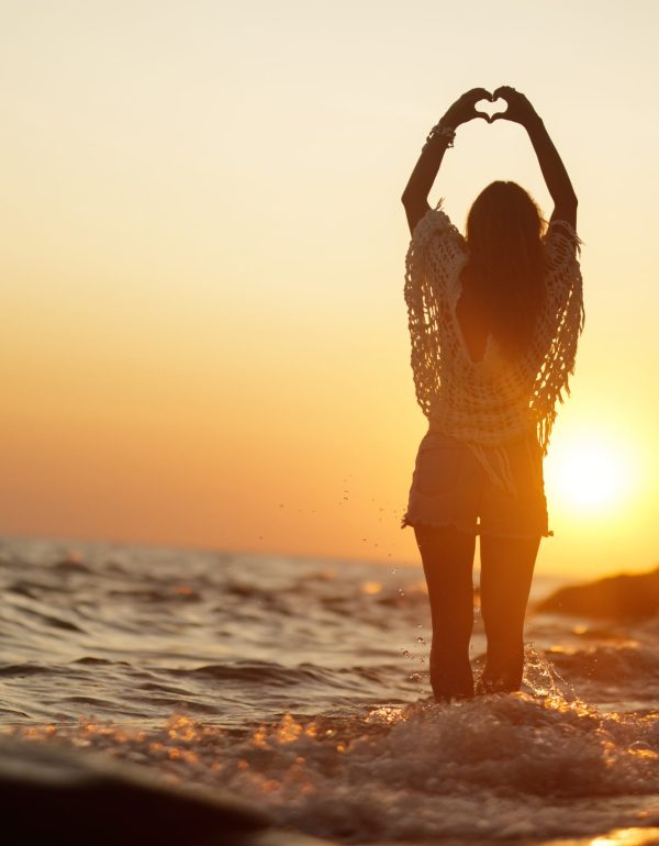Back view of woman standing in the water and making heart shape during summer sunset. Copy space.